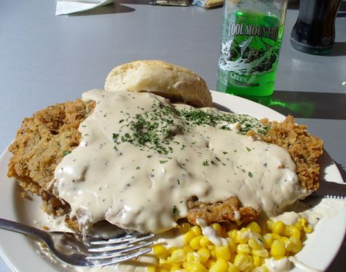 A plate of fried chicken smothered in creamy gravy, served with corn and a biscuit, alongside a green soda.