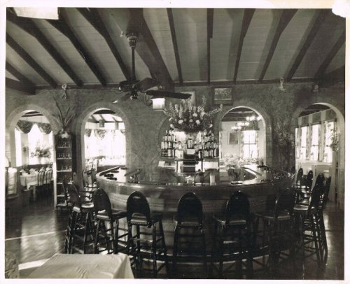 A vintage black-and-white photo of a curved bar with high stools, surrounded by arched windows and decorative plants.