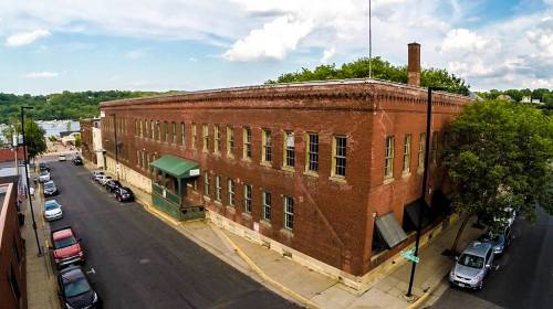Historic brick building on a street, surrounded by trees and parked cars, with a cloudy sky in the background.