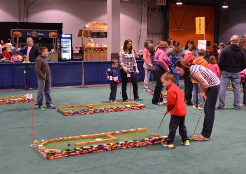 Children and adults play mini-golf at a lively indoor event, with colorful obstacles and a green carpeted floor.