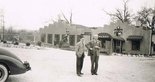 Two men stand outside a building labeled "Basso," with a vintage car parked nearby and trees in the background.