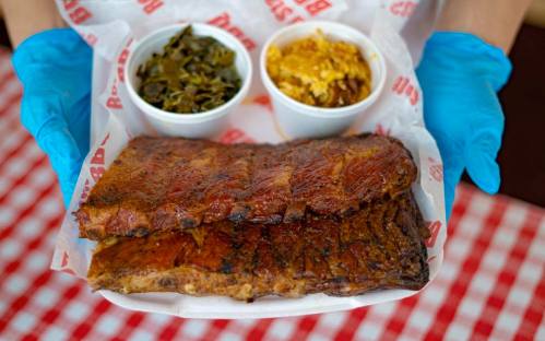 A person in blue gloves holds a tray with two ribs and two sides of collard greens and macaroni and cheese.