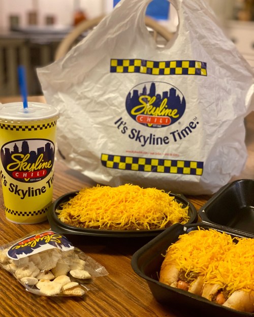 A takeout meal from Skyline Chili featuring two trays of chili cheese coneys, a drink, and a bag of snacks.