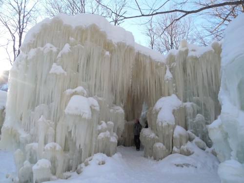A person stands inside a stunning ice cave with towering icicles and snow-covered walls, surrounded by winter scenery.