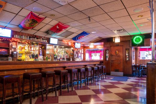 A cozy bar interior with a wooden counter, stools, colorful flags, and a well-stocked bar illuminated by soft lights.