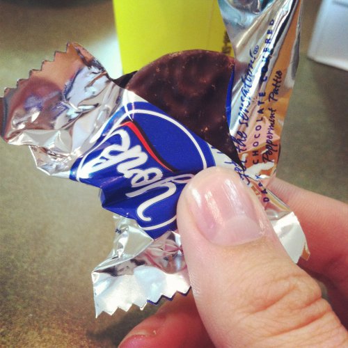 A hand holding a chocolate-covered peppermint patty partially unwrapped from its foil packaging.