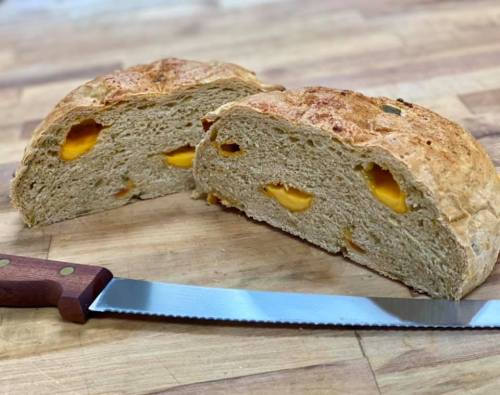 A loaf of bread with chunks of cheese inside, sliced in half, resting on a wooden surface next to a serrated knife.