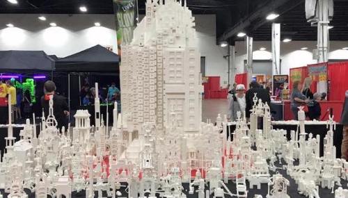 A large, intricate white building model surrounded by smaller structures, displayed at an event with colorful booths in the background.
