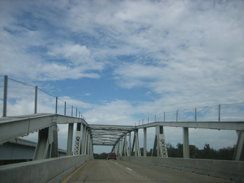A view of a metal bridge with a clear blue sky and scattered clouds above, seen from a vehicle on the road.