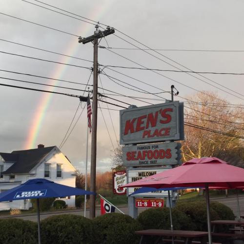 A seafood restaurant sign, "Ken's Place," with a rainbow in the background and colorful umbrellas in the foreground.