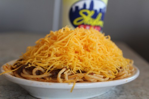A plate of spaghetti topped with a generous mound of shredded cheddar cheese, with a drink in the background.