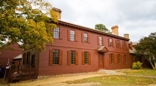 A historic red wooden house with multiple windows and a front door, surrounded by trees and autumn foliage.