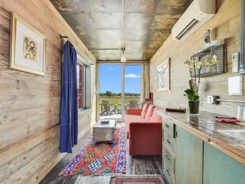 Cozy interior of a small cabin with a red sofa, wooden walls, and a view of the outdoors through large windows.