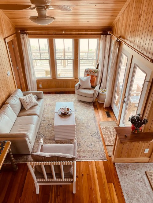 Cozy living room with wooden walls, a white sofa, armchair, coffee table, and large windows letting in natural light.