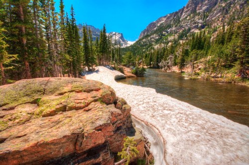 A scenic view of a river flowing through a forested area, with snow patches and mountains in the background.