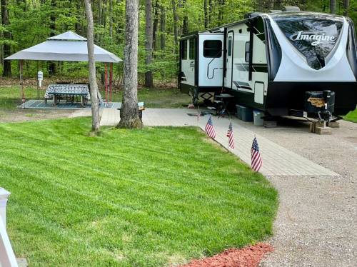 A camper parked in a wooded area with a gazebo and lawn, featuring small American flags along the pathway.