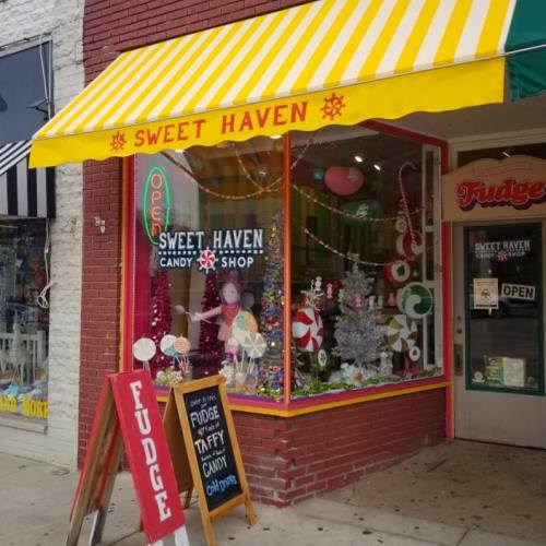 Colorful storefront of Sweet Haven Candy Shop, featuring a bright yellow awning and festive decorations in the window.