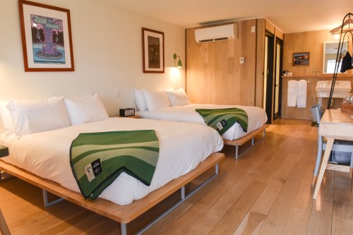 A cozy hotel room featuring two beds with green throws, wooden floors, and modern decor.
