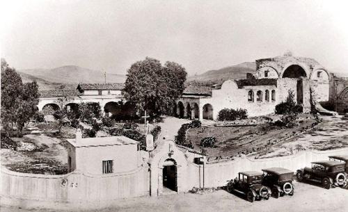 Historic black-and-white photo of a mission complex with arches, gardens, and vintage cars parked outside.