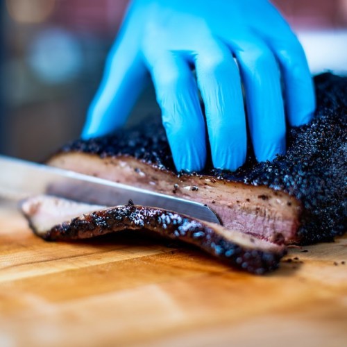 A gloved hand slices into a smoked brisket on a wooden cutting board.