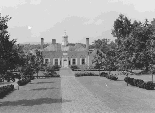 Historic building with a central entrance, surrounded by trees and a paved pathway leading to it. Black and white photo.