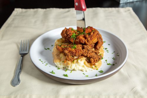 A plate of fried chicken on creamy mashed potatoes, topped with gravy and garnished with parsley, with a knife inserted.