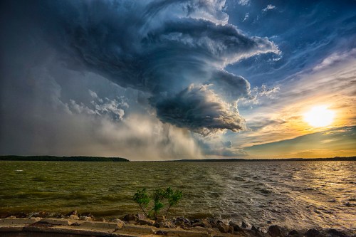 Dramatic storm clouds gather over a lake at sunset, with dark skies contrasting against the warm horizon.