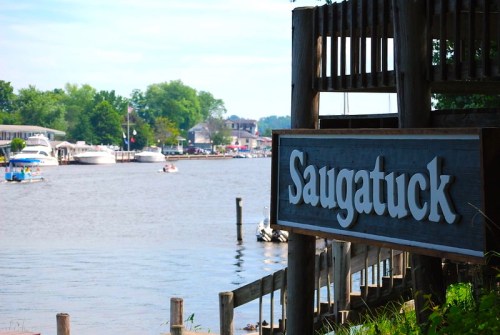 Sign for Saugatuck by a river with boats and trees in the background on a sunny day.