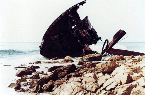A shipwreck partially submerged on rocky shore, with waves lapping at the rusted hull under a clear sky.