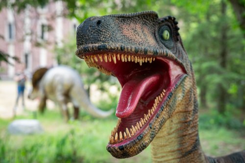 A close-up of a dinosaur statue with an open mouth, set in a lush green environment with another dinosaur in the background.