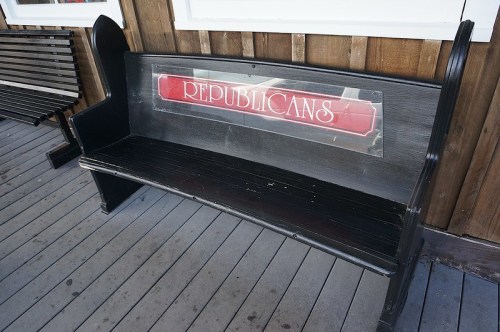 A black wooden bench with a sign that reads "REPUBLICANS" on the backrest, placed on a wooden deck.