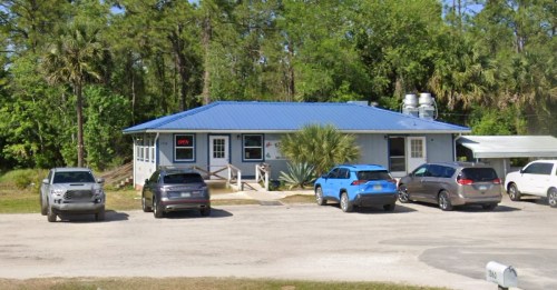 A blue-roofed building surrounded by trees, with several parked cars in front and a sign indicating it's open.