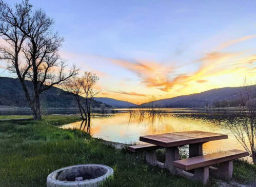 A serene lakeside scene at sunset, featuring a picnic table, trees, and colorful clouds reflecting on the water.