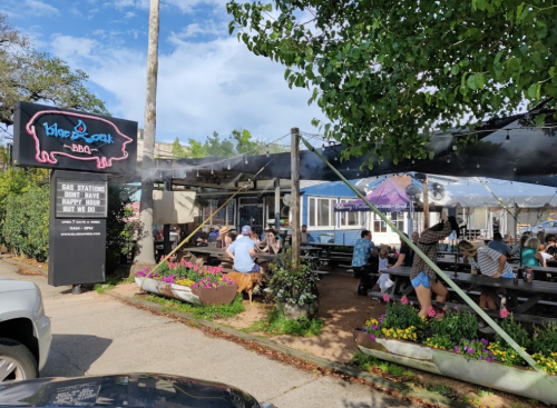 Outdoor dining area of a BBQ restaurant with patrons seated under a shaded canopy, vibrant signage visible.