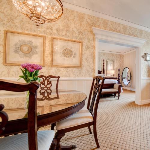 Elegant dining area with a round table, floral centerpiece, and a view into a beautifully decorated bedroom.