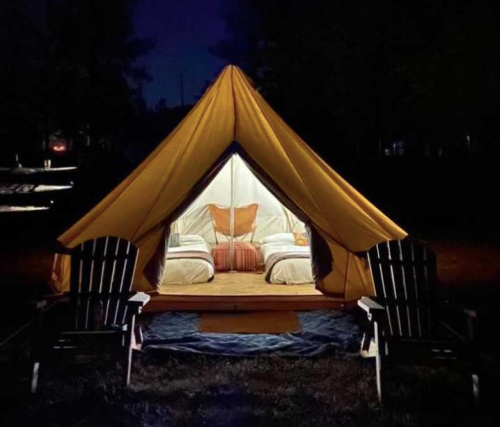 A cozy glamping tent at night, featuring two beds inside and two wooden chairs outside.