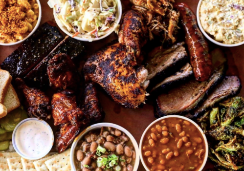 A vibrant spread of barbecue meats, sides, and bread, including ribs, chicken, sausage, coleslaw, and baked beans.