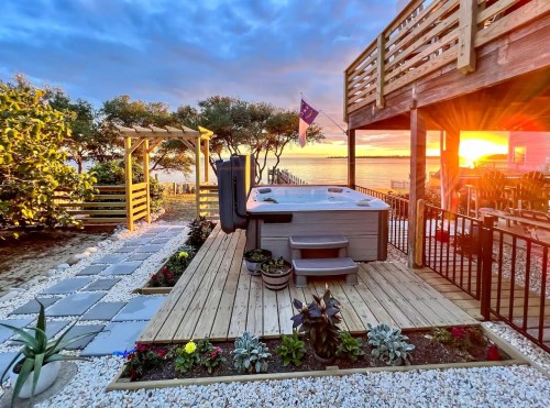 A serene outdoor space featuring a hot tub, wooden deck, and sunset over the water, surrounded by plants and flowers.