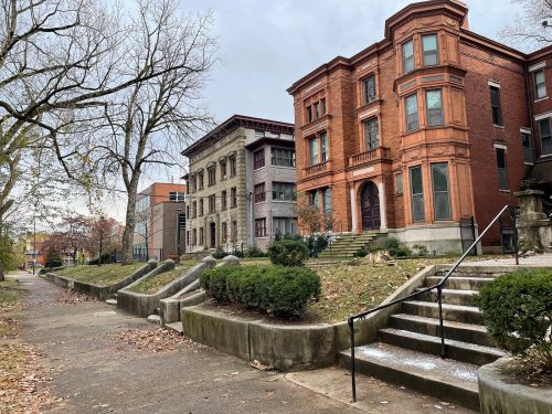 Historic brick buildings line a tree-lined street, with steps leading up to their entrances and a cloudy sky above.