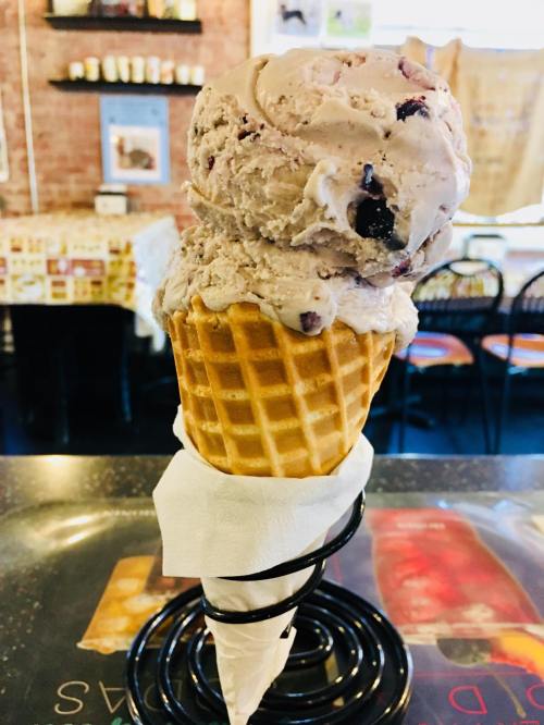 A waffle cone holds two scoops of blueberry ice cream, resting on a black wire holder in a cozy ice cream shop.