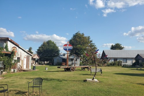 A rustic outdoor area with buildings, sculptures, and green grass under a blue sky with scattered clouds.