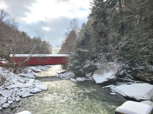 A snowy landscape featuring a red covered bridge over a flowing river, surrounded by trees and rocky banks.