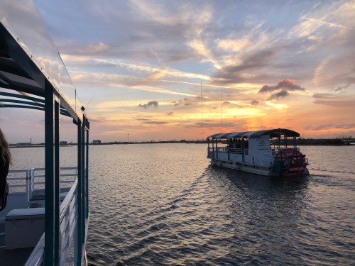 A boat sails on calm waters at sunset, with vibrant colors in the sky and another boat in the distance.