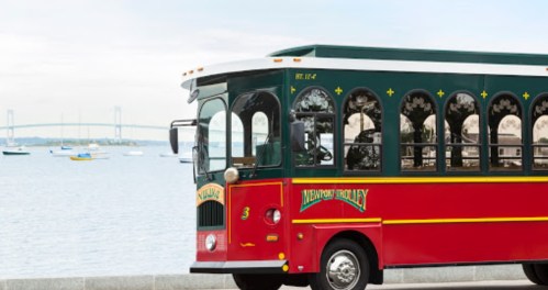 A red and green trolley bus parked by a waterfront with boats and a bridge in the background.