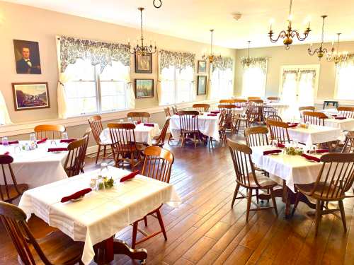 A cozy restaurant interior with wooden chairs, white tablecloths, and large windows, featuring elegant chandeliers and artwork.
