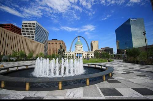 A fountain in a plaza with a statue, surrounded by modern buildings and a blue sky with clouds.