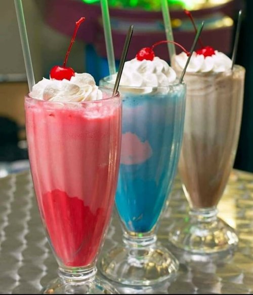 Three colorful milkshakes in tall glasses, topped with whipped cream and cherries, on a shiny silver table.