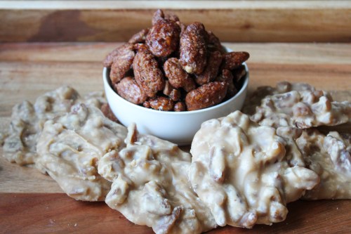 A bowl of spiced nuts sits atop a wooden surface, surrounded by clusters of creamy pralines.