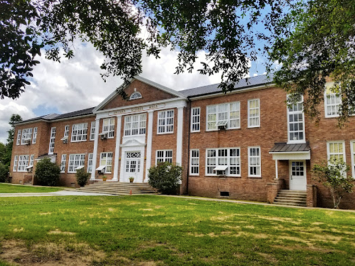 A large brick building with multiple windows, a central entrance, and a grassy area in front, under a partly cloudy sky.