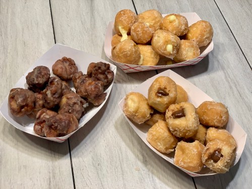 Three trays of assorted donuts: glazed, filled, and cake-style, arranged on a wooden table.